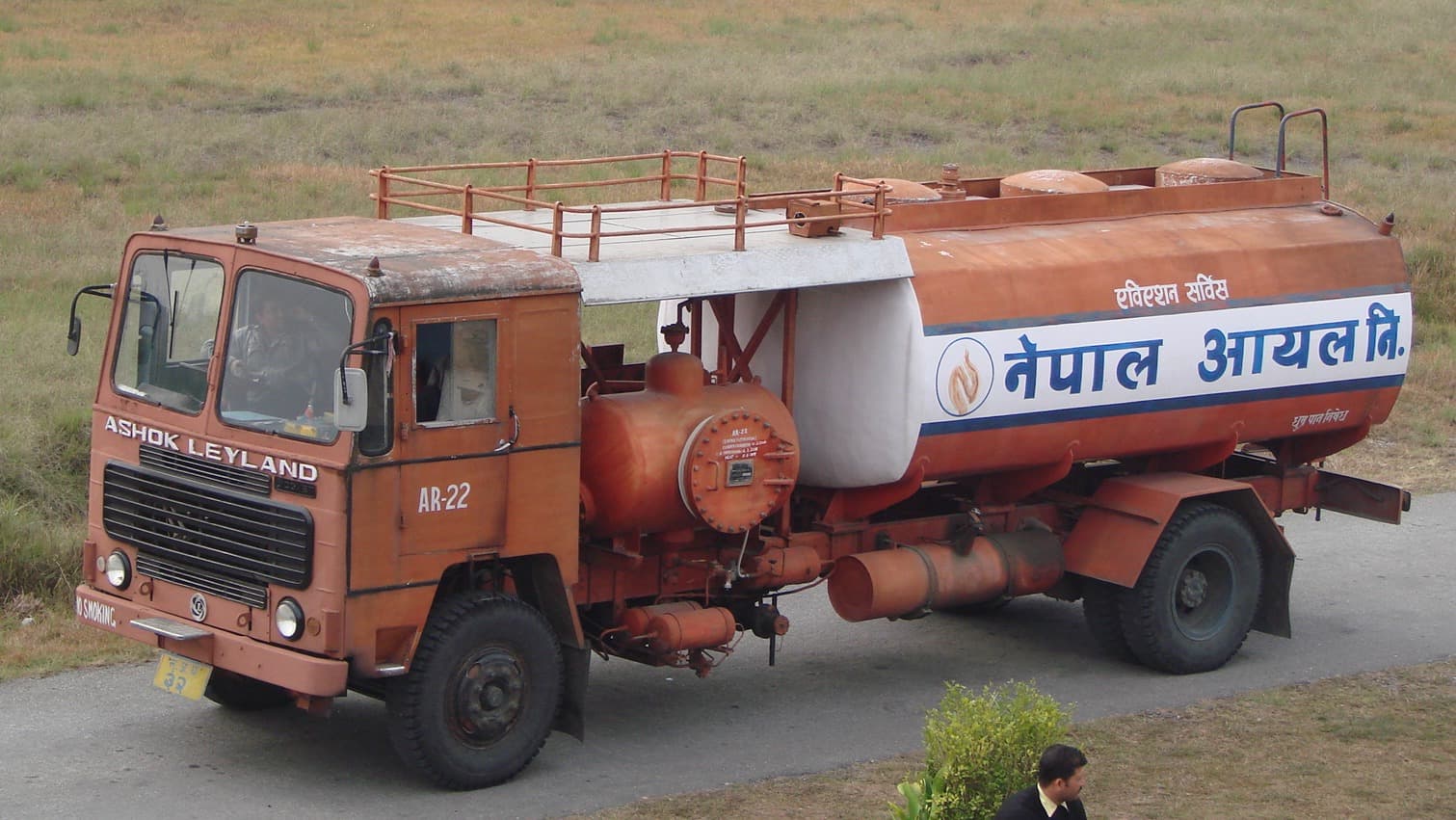 A Ashok Leyland tanker truck, painted in orange with a white stripe and the Nepali text for "Nepal Oil Corporation," is stationary on a paved area.