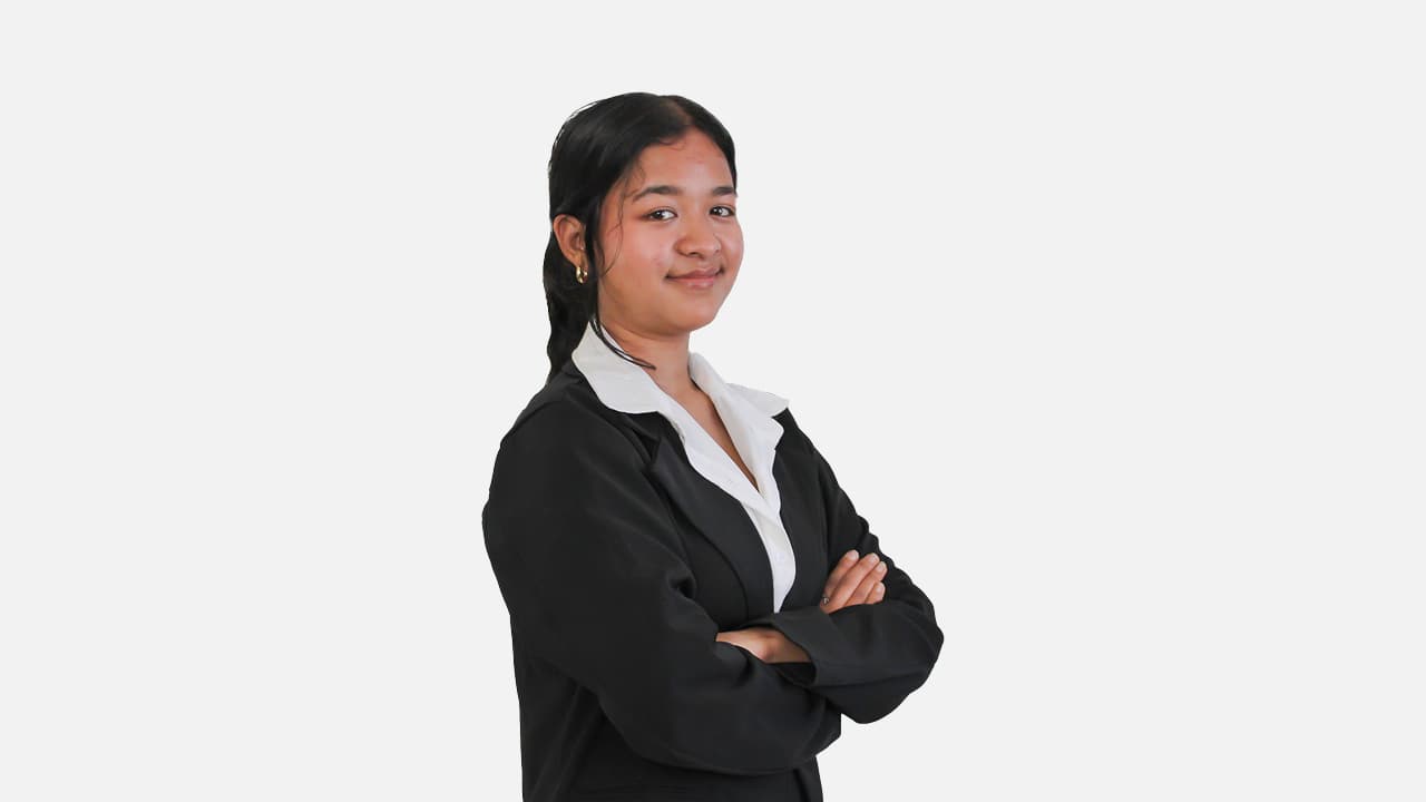 A professional headshot of a young woman with dark hair pulled back, wearing a black blazer over a white shirt, smiling slightly with her arms crossed against a plain white background.