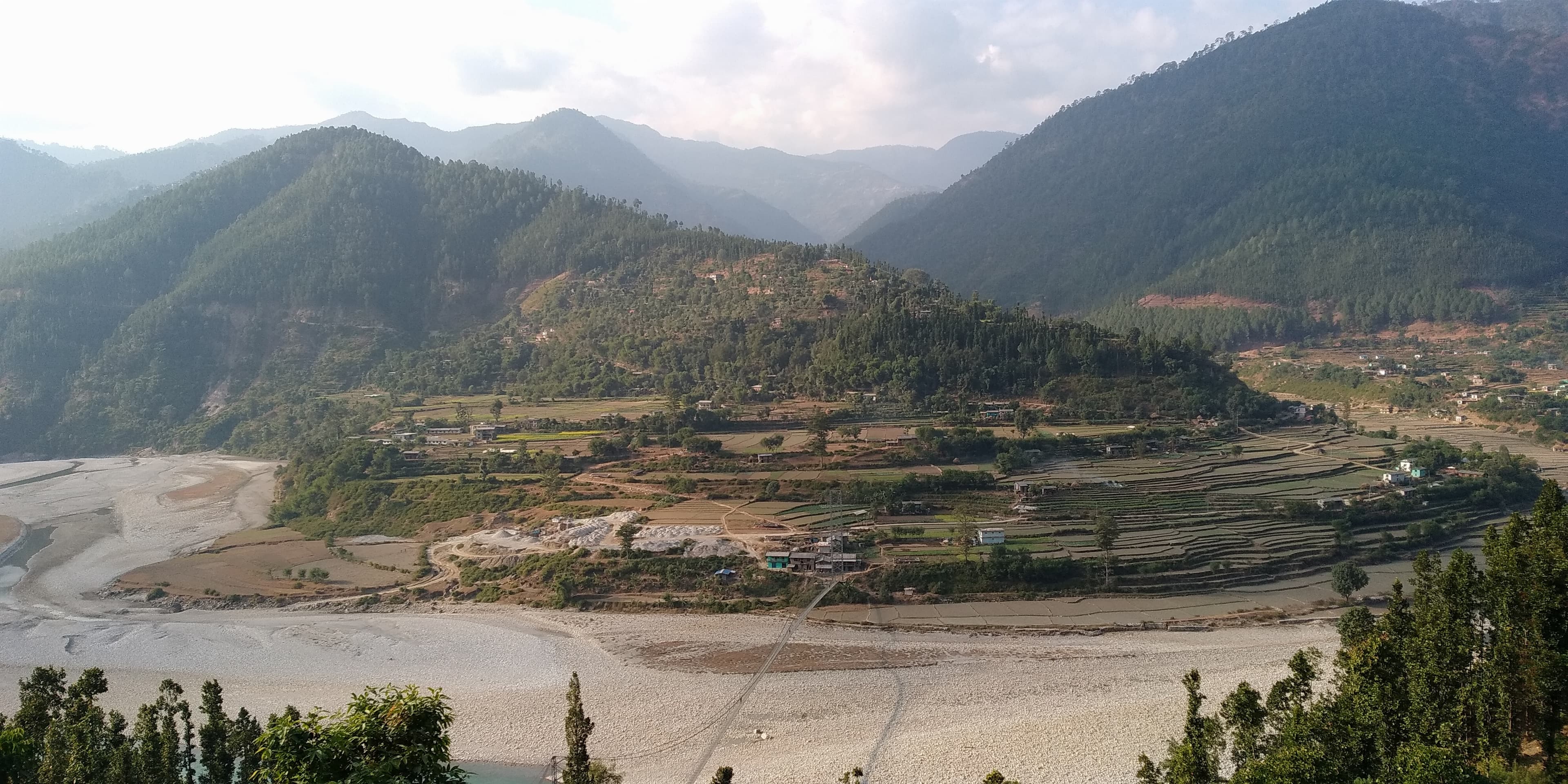 A wide shot of Kalegaun, Jajarkot valley with a dry riverbed, terraced fields on rising slopes dotted with houses, and hazy mountains under a partly cloudy sky.