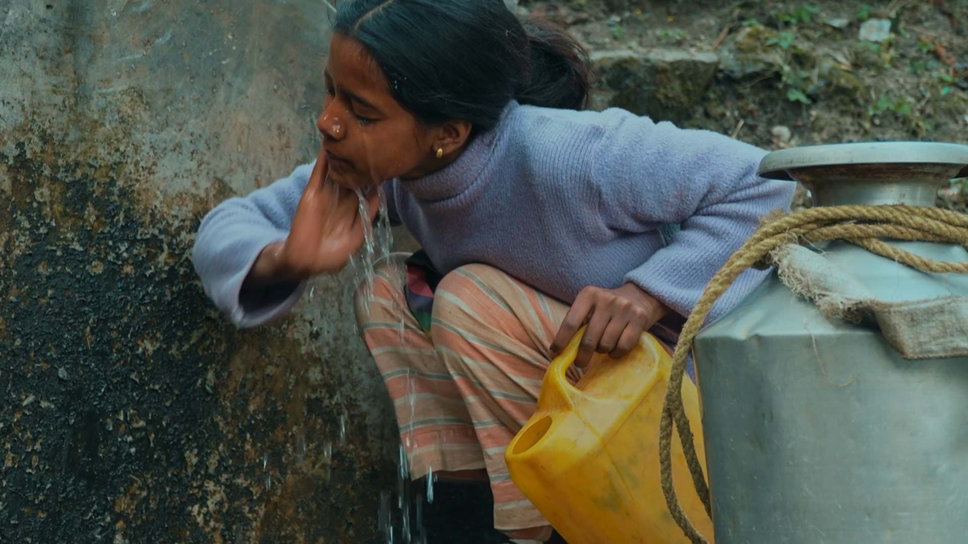 A young girl in a purple sweater and striped pants washes her face with water flowing from a wall spout while holding a yellow container. A large metal water vessel with a rope tied around it sits beside her.