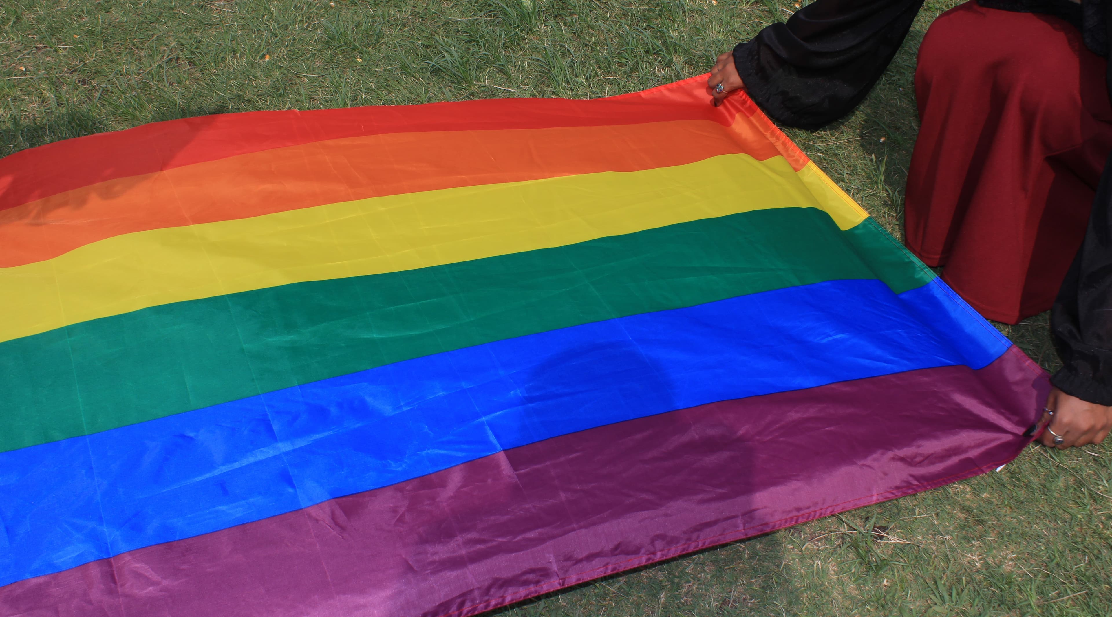 A person's hands are visible on the edges of a large, unfolded rainbow flag lying flat on green grass, with part of their red clothing also visible.