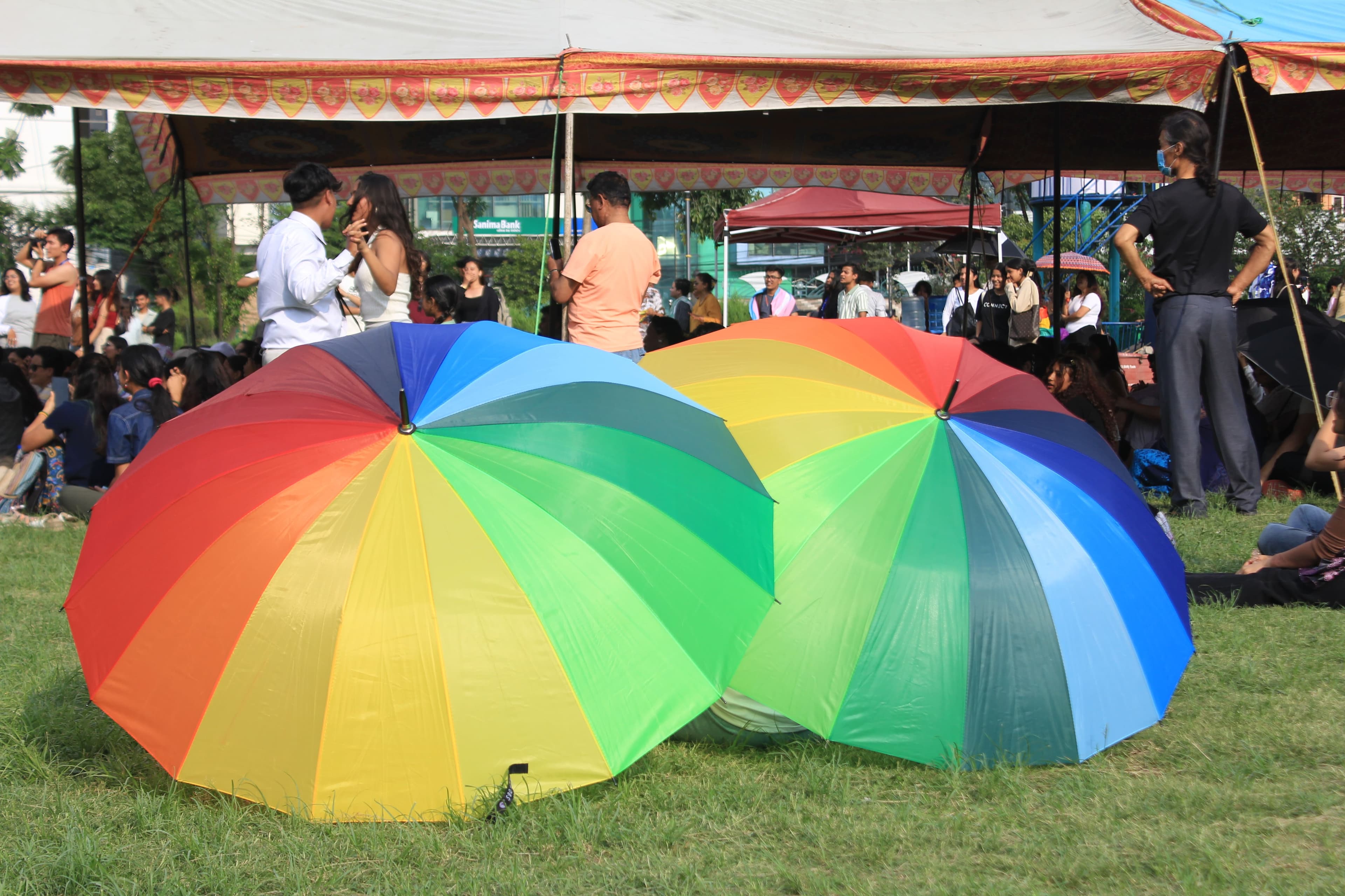 Two large, open rainbow-colored umbrellas rest on green grass, with people gathered under tents in the background.