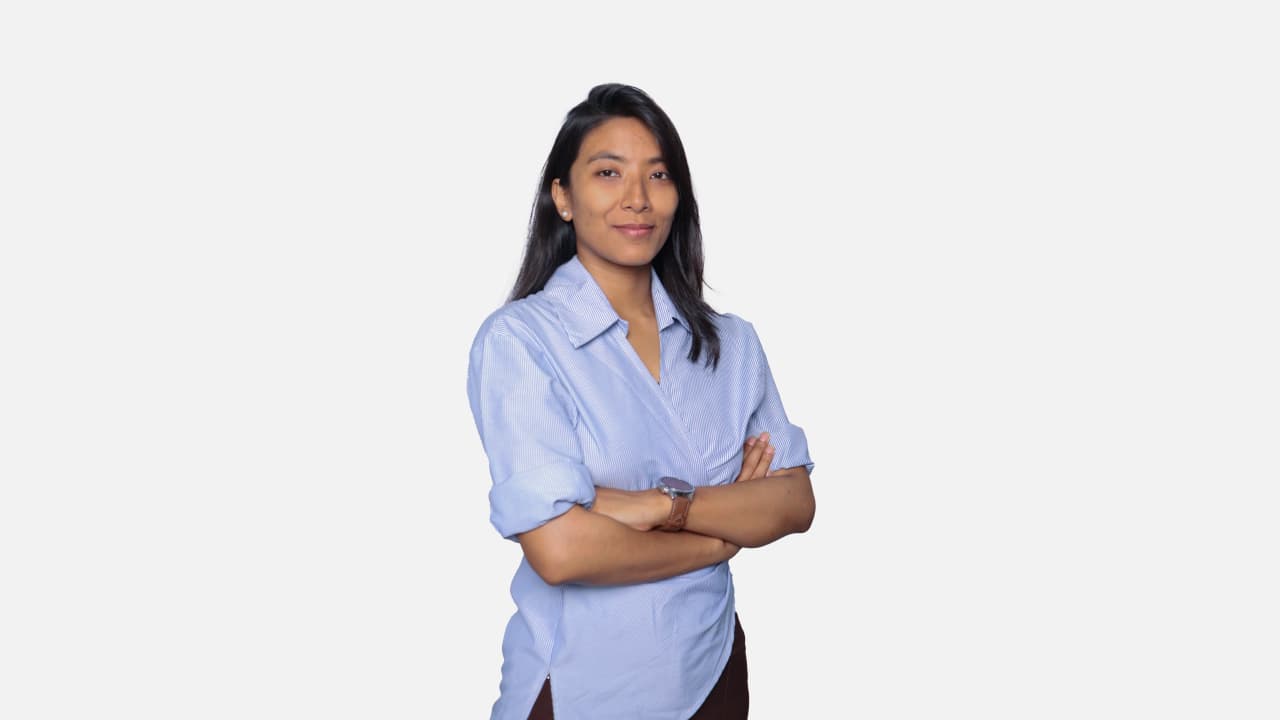 A professional headshot of a young woman with dark hair and a light complexion, wearing a blue collared shirt with her arms crossed, smiling slightly against a plain white background.