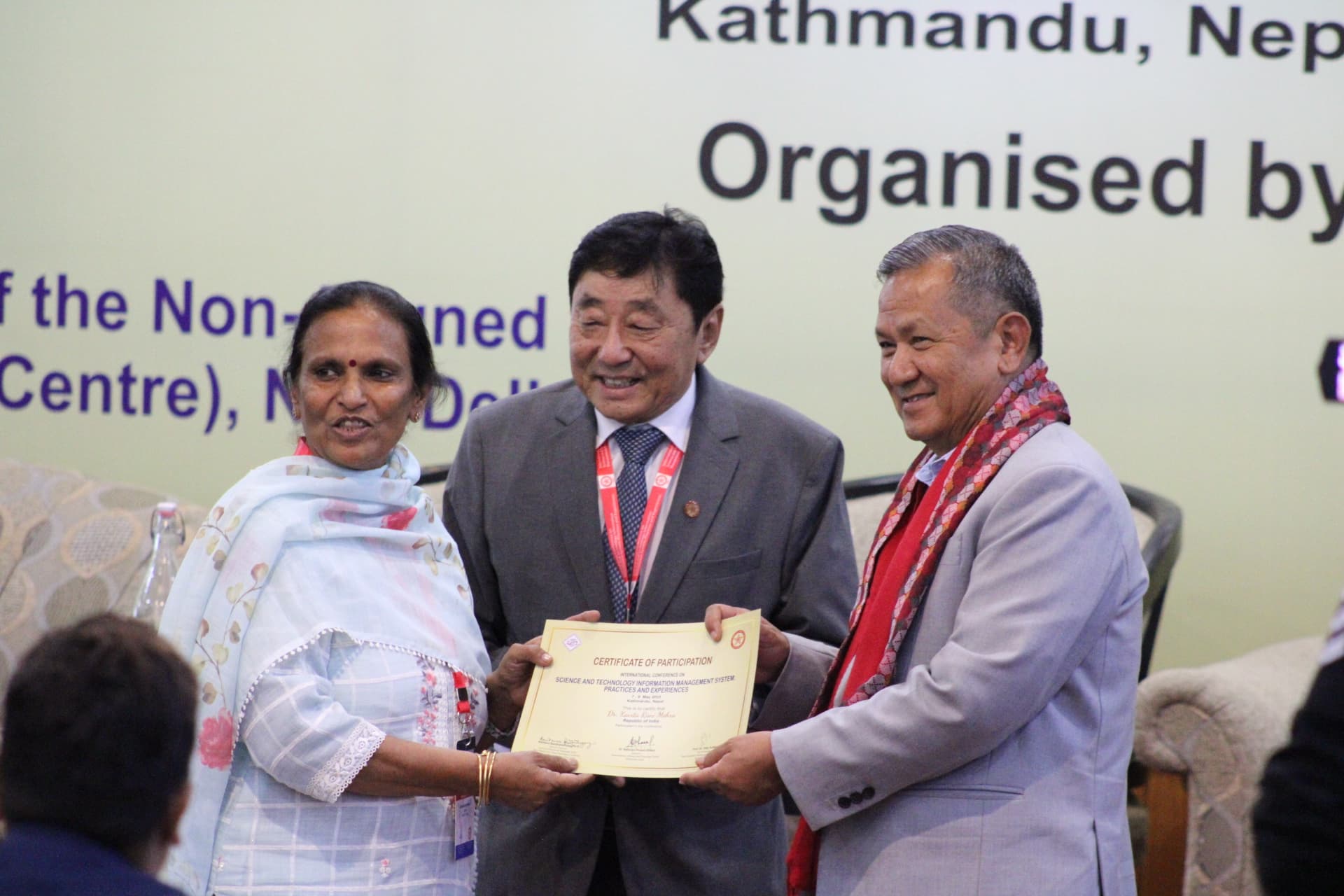 An older woman in a light blue patterned outfit and a man in a dark suit with a red tie present a certificate of participation to a man in a grey suit and red patterned scarf, all smiling on a stage.