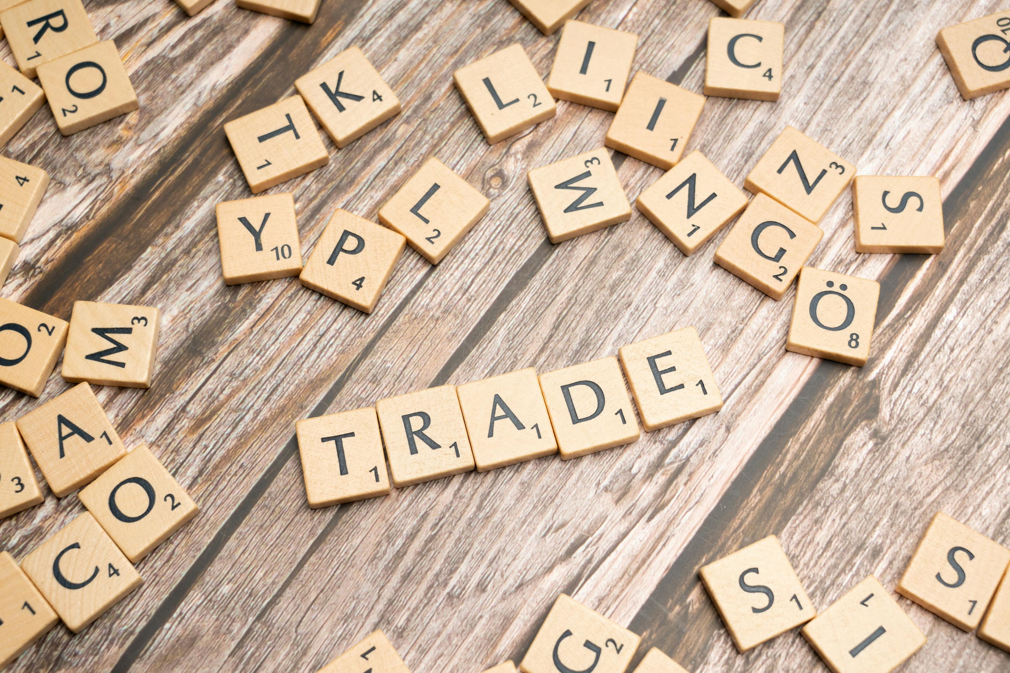 The word "TRADE" spelled in wooden tiles sits amidst scattered letters on a textured wood surface.